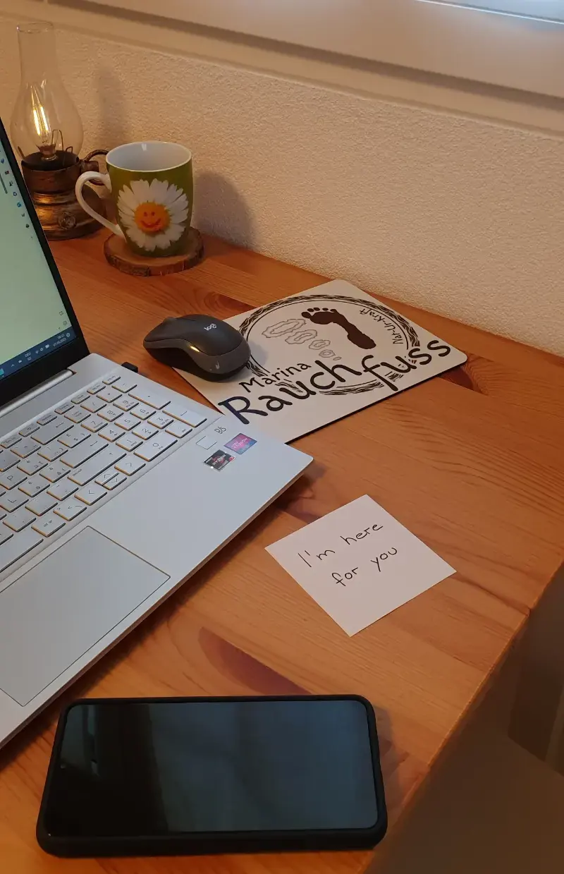 Der Arbeitstisch von Marina Rauchfuss mit dem Laptop, der Mausmatte mit dem Logo, eine Tasse, ein Handy und ein Postit auf dem steht: I'm here for you