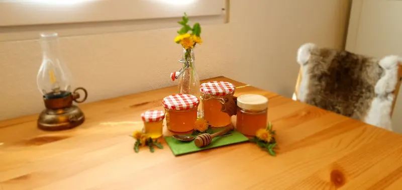 Ein Holztisch mit 4 verschieden grössen von Einmachgläsern dekoriert mit einem Honiglöffel und frischem Löwenzahn