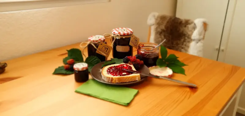 Ein Holztisch mit 4 verschiedenen grössen von Einmachgläsern, ein Teller mit einem Brombeerkonfitürebrot und dekoriet mit frischen Brombeeren und deren Blätter