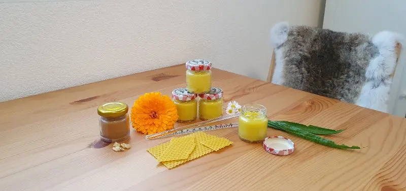Ein Holztisch mit 4 Einmachgläsern von Ringelblumensalbe, dekoriert mit frischen Ringelblumen, Bienenwachs und Salbenutensilien
