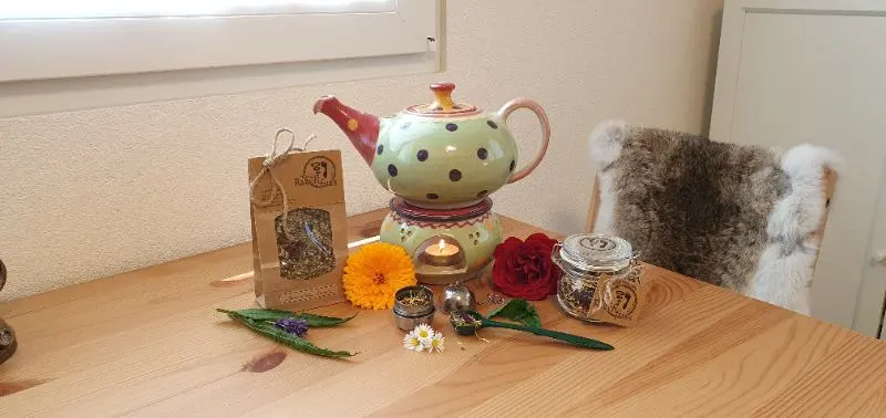 Ein Holztisch mit einer Teekanne, einem Teebeutel gefühlt mit einer Teemischung, ein Einmachglas gefühlt mit Tee, ein Teelöffel und ein Teesieb, dekoriert mit Blüten und Kräutern